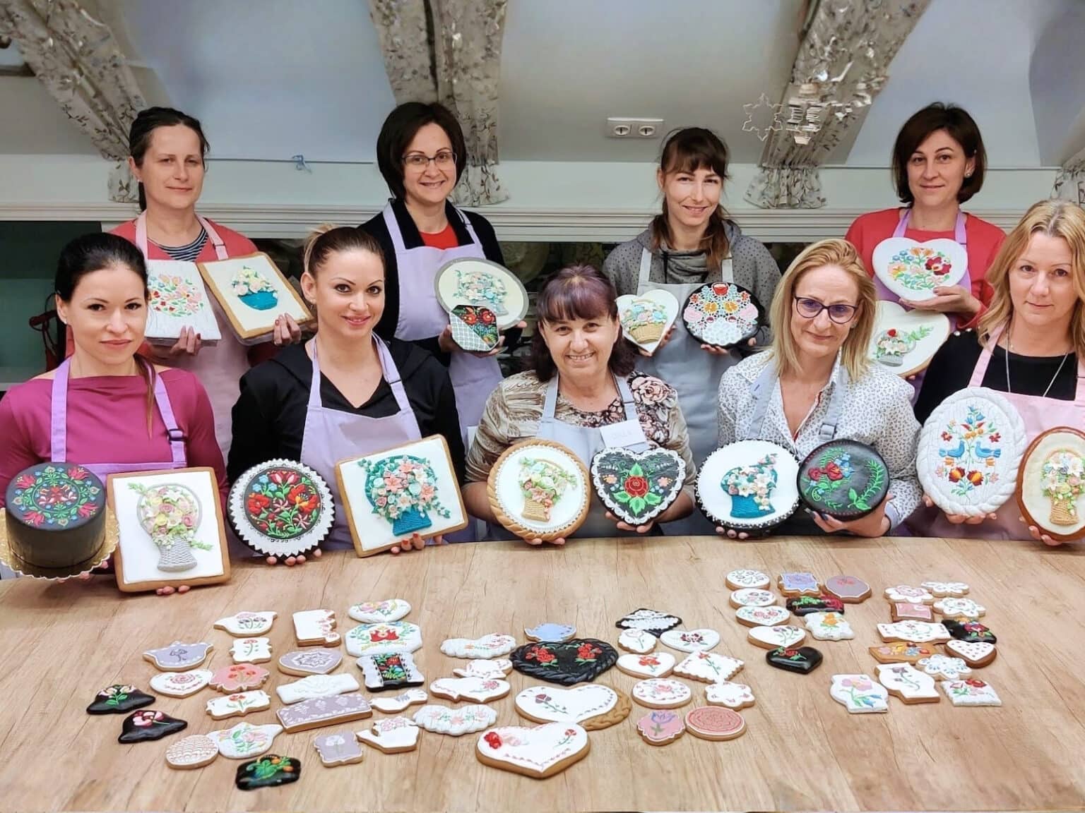 Ékes-Édes Stúdió csoportkép a tanfolyam végén mosolygó emberek és rengeteg elkészült szép cukormázzal díszített mézeskalács az előtérben.