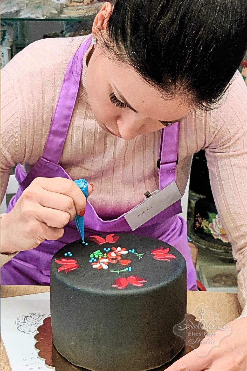 Nő tortát díszít színes cukormázas tedhnikával és részletes népi motívumokkal, közben a dekorációra koncentrálva – Ékes-Édes Stúdió, mézeskalács díszítő tanfolyam