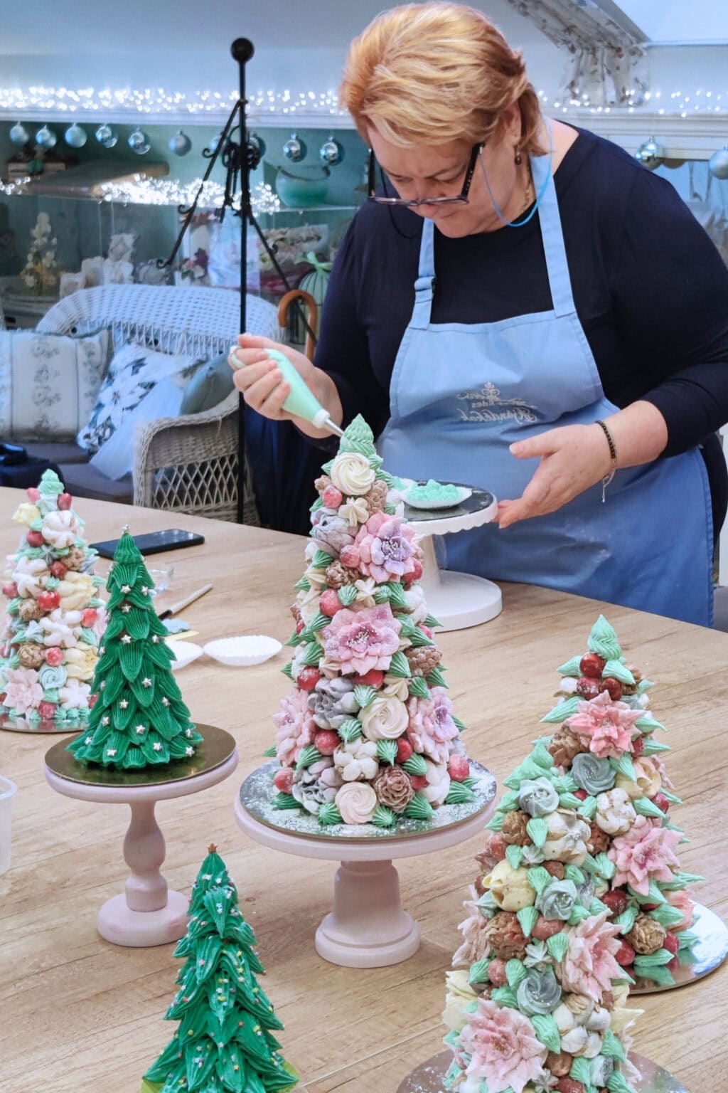 Ékes-Édes Stúdió kreatív cukrászati műhelyében: az asztalon előtérben az elkészült pillecukor elegáns fenyők és karácsonyfák, a háttérben egy tanuló habzsákkal figurákat készít.
