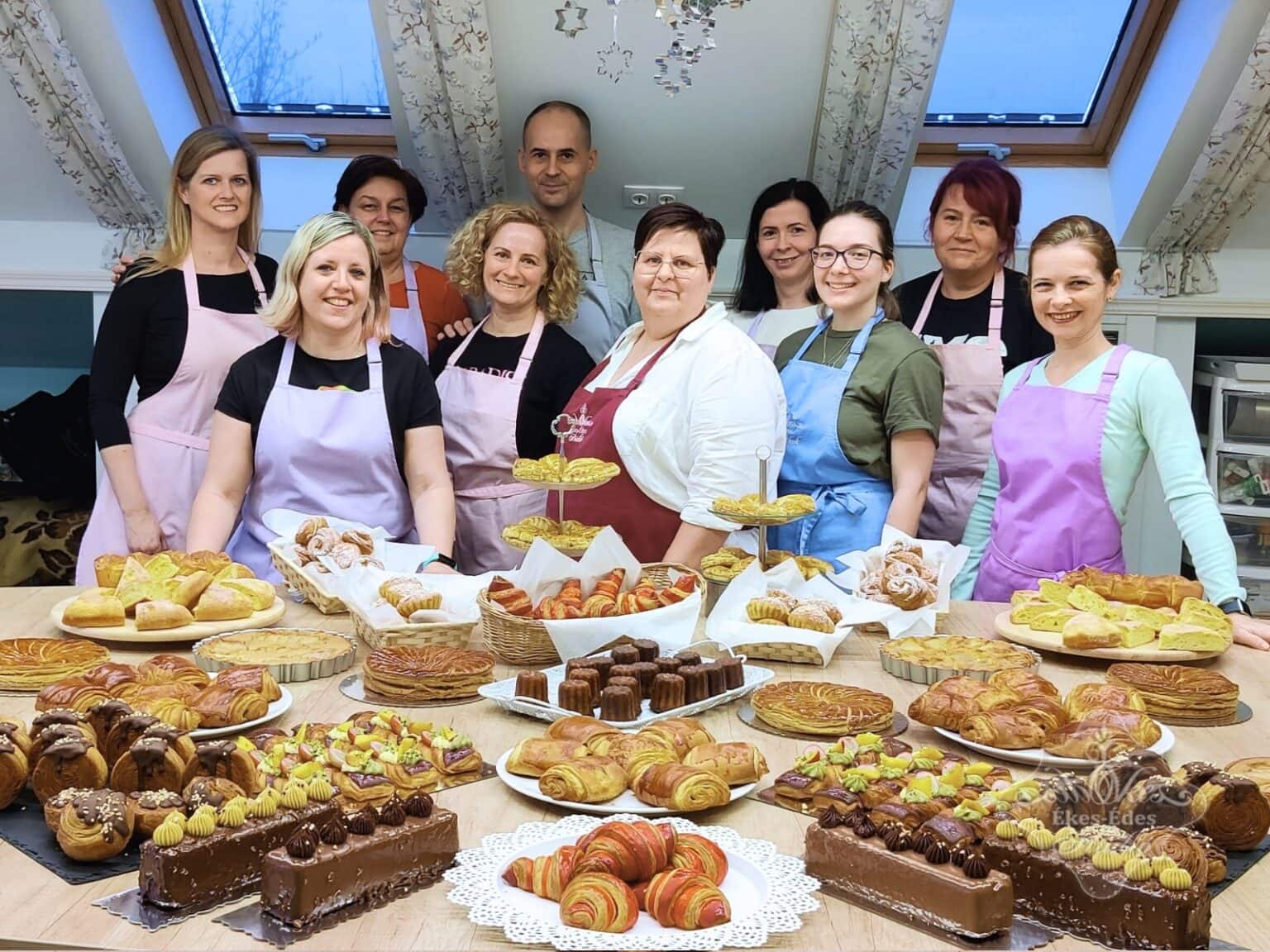 Elégedett emberek csoportképen a tanfolyam végén az elkészült sokféle süteménnyel az előtérben.