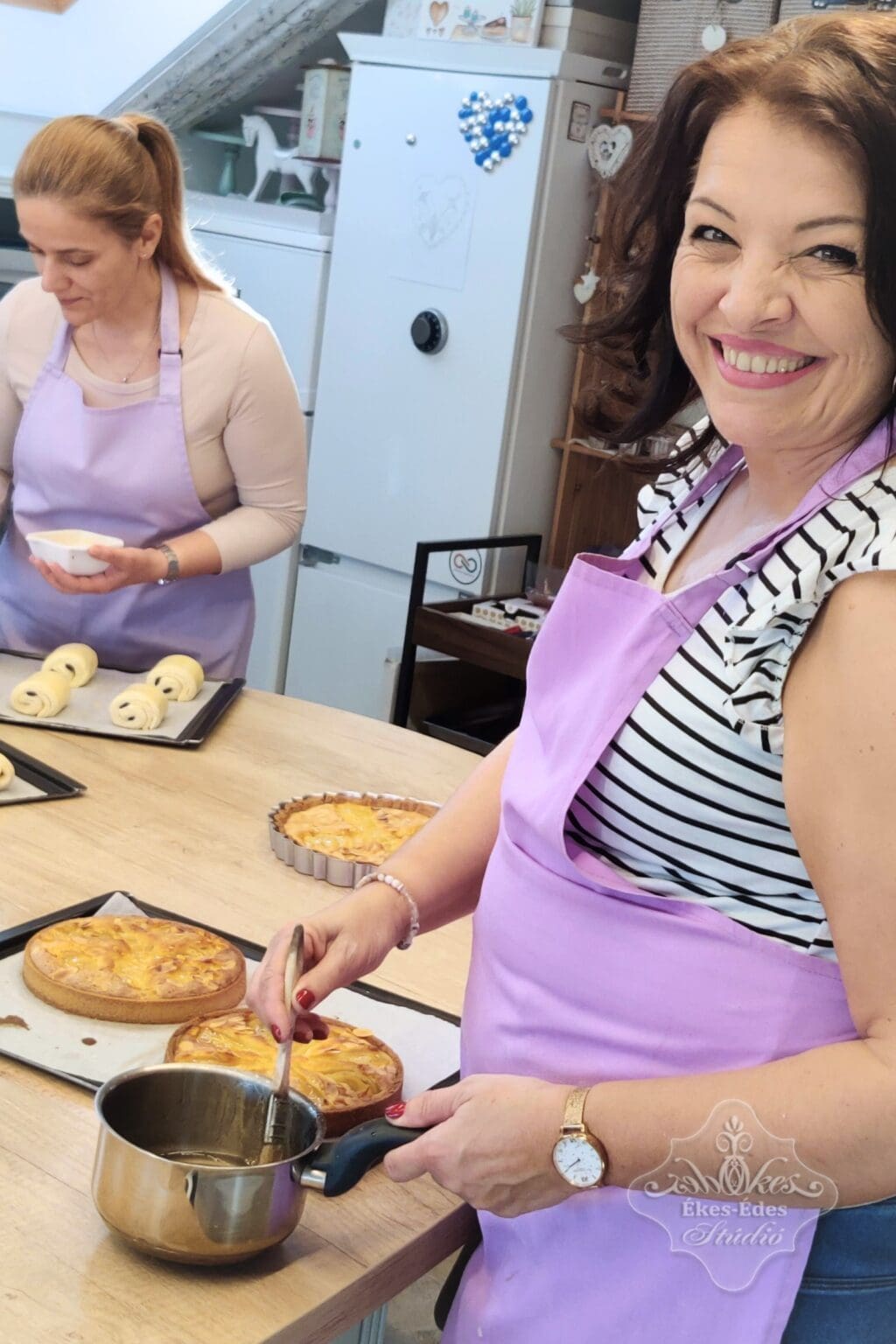 A kép bemutatja, ahogy egy nő süteményt készít, miközben másik résztvevő is dolgozik a háttérben, barátságos és kreatív környezetben az Ékes-Édes Stúdióban, barátságos környezetben, szakértő oktatókkal.