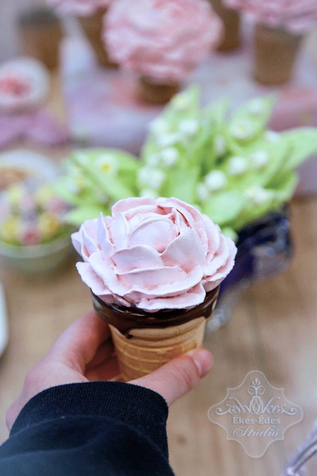 Csokoládés fagylalt tölcsér rózsaszín pillecukor rózsa díszítéssel, háttérben virágok és édességek.