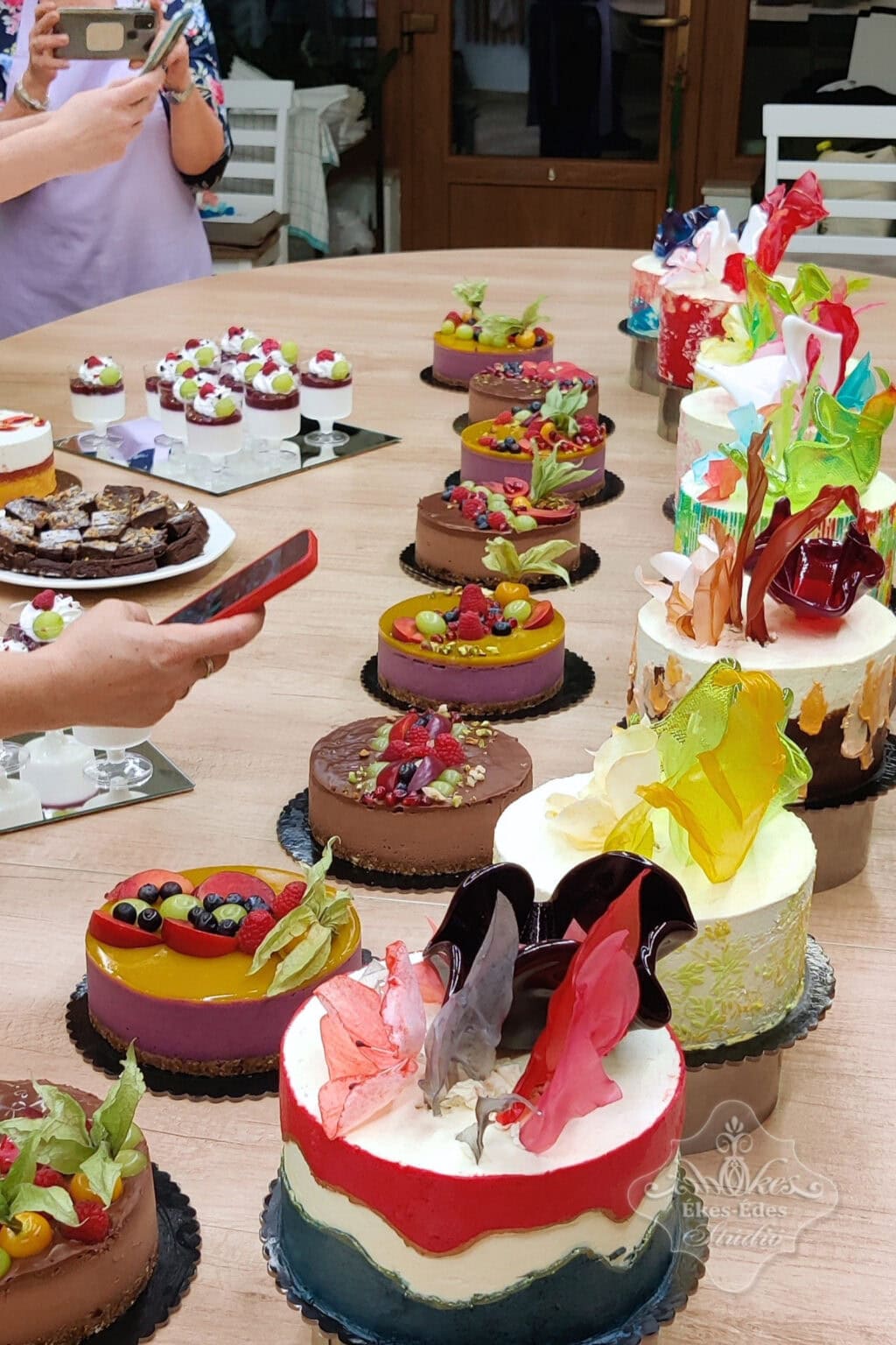 Tanfolyam végén a színes dísztett és nyers vegán torták sora. Bal szélen a résztvevők telefonjaikkal a kezekben épp fotókat készítenek a gyönyörű látványról.