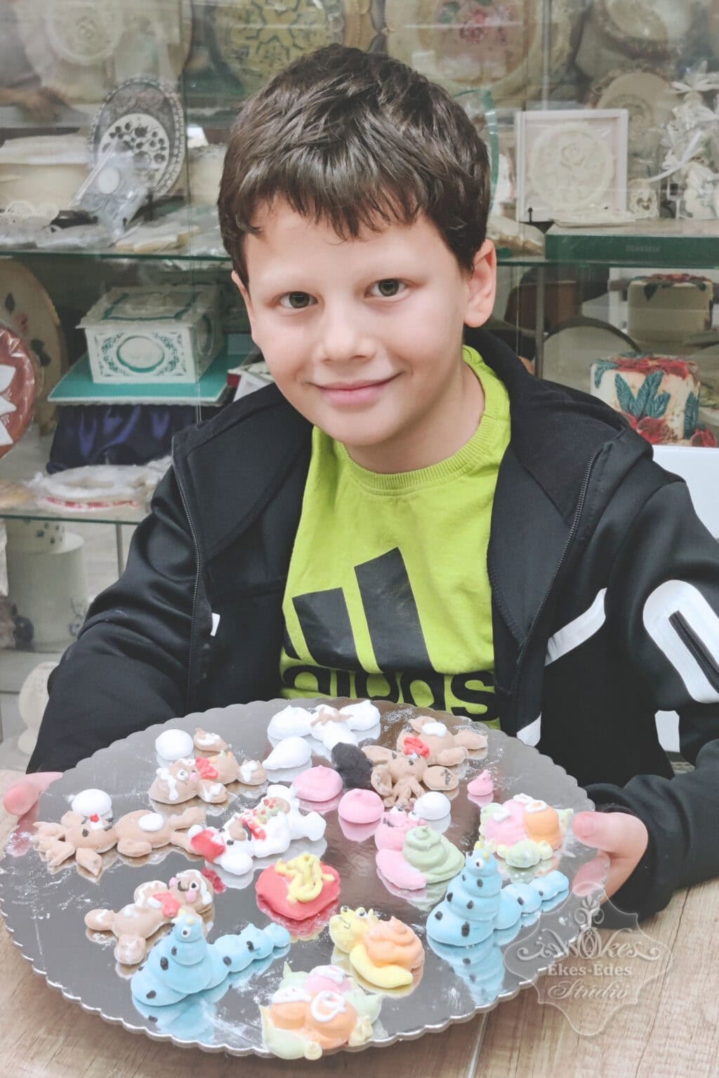 Ékes-Édes Stúdió Pillecukor tanfolyamának legfiatalabb résztvevője, egy mosolygós kisfiú, tálcán mutatja az elkészített színes kedves, duci figuráit.